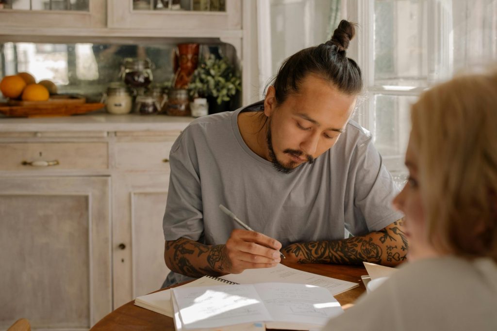 Tattooed young adults studying and taking notes at a table in a warmly lit room.